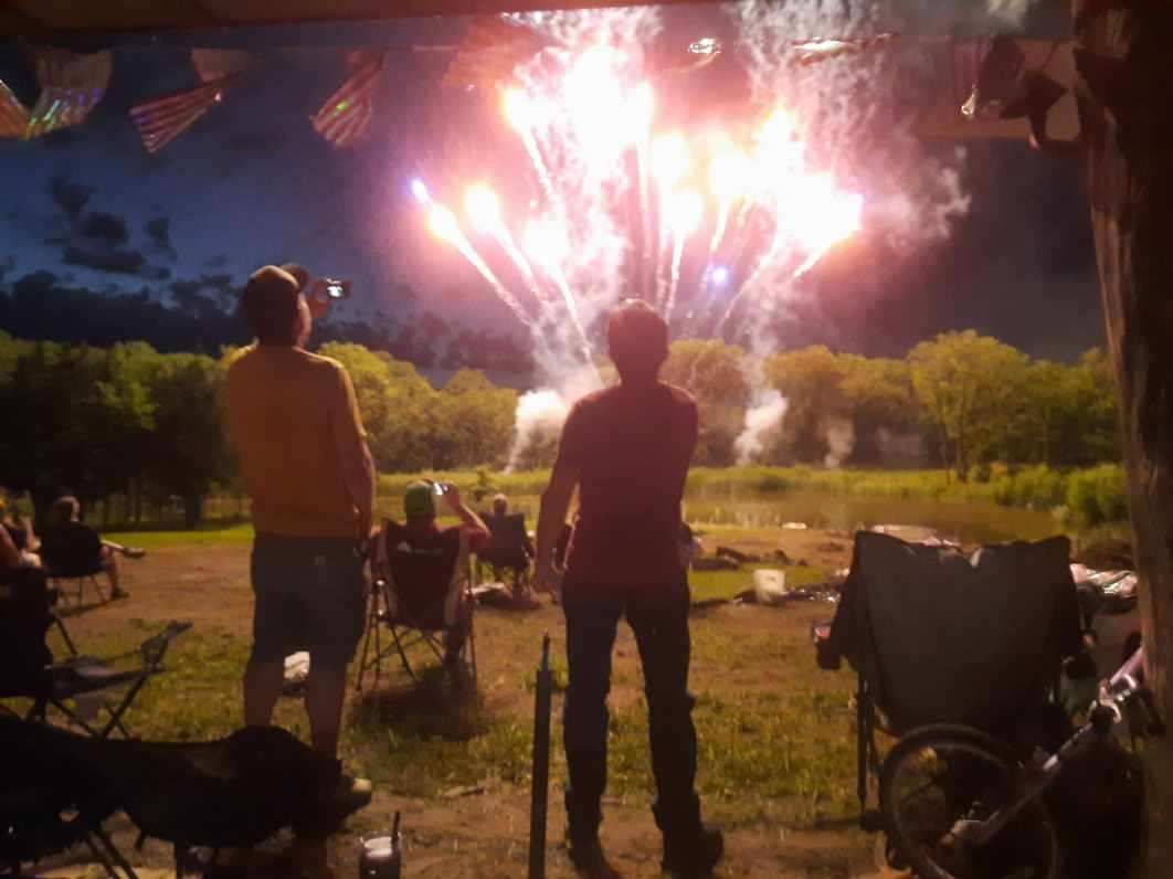 Neighbors gathered at dusk to watch fireworks over a pond, lawn chairs and bunting around them
